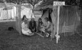 Missionsläger 10 augusti 1967

En vuxen kvinna sitter utanför ingången till ett tält tillsammans med tre stycken ungdomar- två flickor och en pojke. Runt tältet löper ett staket sammanfogat av pinnar. Hon visar dem en lång pinne fylld med hål. En afrikansk skulptur står lutad mot hennes knä. En skylt står vid sidan om hyddan med texten 