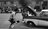 Mosåsflicka, Brandkårsuppvisning, Harar, Transportkillar 27 maj 1967

Tre män i maskeradkläder är i färd med att släcka en brand i motorhuven på en bil under en branduppvisning. Mannen närmast på bilden är klädd i skjorta, slips, shorts, strumpor, skor och med en hatt på huvudet. Mannen i mitten är klädd i klänning och hatt. Han försöker släcka elden i motorhuven genom att vifta med klänningen! Mannen till höger är klädd i skjorta och hatt. Publik åser det hela i bakgrunden.