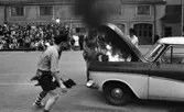 Mosåsflicka, Brandkårsuppvisning, Harar, Transportkillar 27 maj 1967

Tre män i maskeradkläder är i färd med att släcka en brand i motorhuven på en bil under en branduppvisning. Mannen närmast på bilden är klädd i skjorta, slips, shorts, strumpor, skor och med en hatt på huvudet. Mannen i mitten är klädd i klänning och hatt. Han försöker släcka elden i motorhuven genom att vifta med klänningen! Mannen till höger är klädd i skjorta och hatt. Publik åser det hela i bakgrunden.
