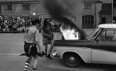 Mosåsflicka, Brandkårsuppvisning, Harar,Transportkillar 27 maj 1967

Tre män i maskeradkläder är i färd med att släcka en brand i motorhuven på en bil under en branduppvisning. Mannen närmast på bilden är klädd i skjorta, slips, shorts, strumpor, skor och med en hatt på huvudet. Mannen i mitten är klädd i klänning och hatt. Han försöker släcka elden i motorhuven genom att vifta med klänningen! Mannen till höger är klädd i skjorta och hatt. Publik åser det hela i bakgrunden.