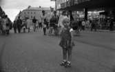 Marknadsafton 16 juni 1967

En liten flicka klädd i en liten klänning, hätta på huvudet, underbyxor, strumpor och sandaler står på gatan i centrala staden Örebro under en marknadsafton. Det är fullt av folk på gatan, däribland mammor med barnvagnar och man ser affärer och byggnader runtomkring.