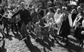 Midsommar, 26 juni 1967

Närbild på barn, kvinnor och män som dansar ringdans under midsommarfirande. Det är flera stycken ringar av dansande människor. Två kvinnor till höger är klädda i folkdräkt. Många män är klädda i kostymer. En mikrofon syns till vänster i bild.