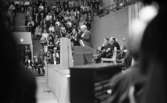 Massmöte 2 maj 1967

Två äldre män klädda i kostymer, skjortor och slipsar står invid varsin mikrofon och talar i den uppe på en scen inne i en arena. Bakom dem sitter ett antal herrar i kostym på stolar. Uppe på läktarna sitter publik och åser det hela. I förgrunden står ett piano.
