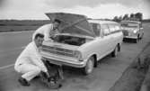 MHF:s vägpatruller 26 juni 1967

Två män i vita arbetsoveraller håller på och lagar en vit bil som står på en väg. En av männen håller på och mekar med verktyg i bilens motorhuv och den andre sitter på huk invid en verktygslåda som står på marken. Bakom den bil de håller på och lagar står deras eget fordon- en bil som har skylten 