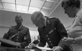 Kungen Museet 28 augusti 1967

Kung Gustav VI Adolf skriver i en stor bok. På ömse sidor om honom står två personer. Den ena närmast i bild är en kvinna klädd i vit dress och glasögon. Den andra personen är en militär. Han bär militärkläder liksom kungen.