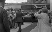 Kungen Museet 28 augusti 1967

Kung Gustav VI Adolf vandrar på röda mattan utanför Örebro Läns Museum klädd i militäruniform och gör honnör mot åskådare som står vid sidan om och betraktar honom. Han har ryggen mot kameran. I bakgrunden står en chaufför och väntar vid en svart bil med uniformsmössan i handen. Han har öppnat bakre bildörren och står beredd att släppa in kungen i bilen. En äldre kvinna klädd i vit kappa står i förgrunden med en kamera och är i färd med att knäppa en bild av kungen.
