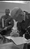 Kungen Museet 28 augusti 1967

Kung Gustav VI Adolf skriver i en bok inne på Örebro Läns Museum. Han har lagt boken på en glasmonter. Han är klädd i militäruniform. Bakom honom står en annan man som också klädd i militäruniform. Han håller i en stor bok.