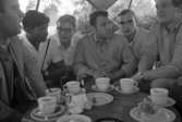 Orubricerad 5 augusti 1967 Travkusk Bengt Nilsson

Sex unga män sitter utomhus kring ett runt bord som har fullt med kaffekoppar, fat och assietter på sig. Fyra stycken av männen är klädda i skjortor, en har T-tröja och en bär jacka.
