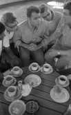 Orubricerad 5 augusti 1967 Travkusk Bengt Nilsson

Fem unga män sitter vid ett runt bord. Fyra av dem är klädda i kortärmade, ljusa skjortor och ljusa långbyxor och den femte har en ljus T-tröja samt ljusa långbyxor. Två av dem bär glasögon. På bordet står kaffekoppar, kaffefat och assietter m.m.