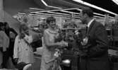 Orubricerad 18 maj 1967

En kostymklädd man skakar hand med en kvinna klädd i kortärmad ljus klänning med krage runt halsen inne i en lokal. Båda har buketter med blommor i händerna. Framför kvinnan står en moped. Till vänster i bakgrunden står tre kvinnor och ler medan de betraktar det handskakande paret.