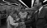 Orubricerad 18 maj 1967

En kostymklädd man skakar hand med en kvinna klädd i en vit kappa inne i en lokal. Hon har en radio i vänster hand med en bukett ovanpå. Hon skakar hand med ovan nämnde man. I bakgrunden står en kvinna klädd i ljus, kortärmad klänning med krage på och håller i en blombukett. En annan kostymklädd man står bredvid henne.
