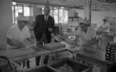 Orubricerad 21 augusti 1967

Tre kvinnor i vita arbetskläder med vita mössor till tillverkar kex på Örebro Kexfabrik. Direktören står mittemellan två kvinnor som jobbar. Han är klädd i mörk kavaj, mörka byxor, vit skjorta och mörk slips. Han håller ett kex i sin vänstra hand. Kvinnorna står invid arbetsbord i metall.