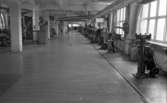 Kembels slutar, Blå stjärnan, 8 juli 1967

En fabrikshall där skotillverkning sker. I bakgrunden syns en man i vit arbetsklädsel. Stora fönster syns till höger i bild.