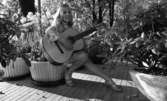 Kommunalmän Wadköping, Bäste amatören 5 juni 1967

En ung flicka sitter och spelar gitarr på kanten av en stor blomkruka. Hon är klädd i en längärmad klänning med volanger och bubbelmönster på. Hon bär pumps med remmar på fötterna. På höger lånfinger bär hon en ring med en pärla i. På vänster vrist bär hon ett armbandsur.