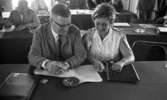 Parti ### (soc.), 18 augusti 1967

I förgrunden sitter en kvinna vid ett bord under ett partimöte. Hon är klädd i en vit, ärmlös blus med kråsmönster på. Hon samtalar med en man i ljus, rutig kostym, vit skjorta och mörk slips. Kvinnan håller i sina glasögon med vänster hand och mannen håller en cigarr i sin högra hand.