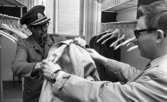 Kurator, Nye generalen 18 maj 1967

En man klädd i skärmmössa, kavaj och som bär glasögon och har en pipa i munnen mottager en rock från en annan man som är klädd i rock, bär glasögon och har ett armbandsur på vänster arm. de befinner sig i en större garderob.