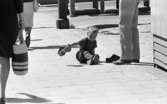 Kvar i stan 26 juli 1967

Ett antal människor befinner sig i centrala stan i Örebro. Närbild på en liten pojke i treårsåldern klädd i svart T-shirt, svarta hängselbyxor, mörka strumpor och mörka sandaler som sitter på gatan och håller i en vuxen mans träsko. Den andra träskon står bredvid honom på marken. En barfota man i vita byxor står bredvid honom. Två kvinnor i klänningar passerar dem.