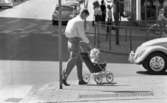 Kvar i stan 26 juli 1967

Ett antal människor befinner sig i centrala stan i Örebro. I förgrunden syns ett litet barn i tvåårsåldern som sitter i en rutig sittvagn. En man i vit skjorta och ljusa byxor kör vagnen. I bakgrunden syns flera personer som går omkring på stan, bilar på gatorna, en cyklist och ett hus.