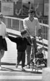 Kvar i stan 26 juli 1967

En familj bestående av mor, far och två små pojkar varav den yngsta pojken sitter i en sittvagn som fadern kör. Mamman håller den större pojken i handen och han håller sin andra hand på broderns vagn. Pojkarna är klädda i T-tröja respektive tröja och byxor, pappan är klädd i vit tröja och ljusa långbyxor, mamman bär vit tröja och kort, mörk kjol.