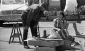 Kvar i stan 26 juli 1967

En kvinna i en kort ärmlös klänning sitter ned nära några lådor på ett torg i centrala Örebro. En kaffetermos står framför henne. En man står invid henne och böjer sig fram och talar med henne. Han är klädd i långärmad mörk tröja, mörka byxor, mörka strumpor och svarta skor. I bakgrunden står en bil parkerad. En kvinna paserar kllädd i vit tröja, vit kjol och vita skor. Hon bär på en handväska.