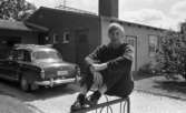 L. Person till USA, Oscaria 28 juni 1967

Närbild på en ung tonårspojke som sitter utanför sin familjs hus. Han är klädd i en mörk långärmad tröja, mörkrutiga byxor, svarta strumpor och svarta skor. En mörk bil står parkerad utanför huset.