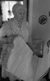 Hobby är avkopplande 25 juli 1967

En gammal dam står med en vit matta i handen. Hon är klädd i en mönstrad kortärmad ljus klänning. På väggen hänger n liten brodyr. En vit stol med mörk dyna står i bakgrunden.