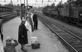 Frövi 2 juni 1967

Resenärer i olika åldrar står på perrongen på Frövi järnvägsstation. I förgrunden syns bl.a. en liten pojke i sjuårsåldern klädd i ljus mössa och ljus jacka. Två äldre damer står i närheten av honom. Den första damen bär en ljus hatt och mörk kappa samt glasögon. En liten mörk resväska med ett paraply i samt en stor vit resväska står på marken mellan damerna. En järnvägstjänsteman stins står lite längre bort på perrongen.