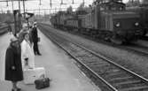 Frövi 2 juni 1967

Resenärer i olika åldrar står på perrongen på Frövi järnvägsstation. Två äldre damer står i förgrunden. Den första damen bär en ljus hatt, mörk kappa, mörka skor samt glasögon. Den andra damen är klädd i vit kappa. En liten mörk resväska med ett paraply i samt en stor vit resväska står på marken mellan damerna. En stins står i bakgrunden och gör honnör. Fler personer syns i bakgrunden. Två tåg syns längre bort.