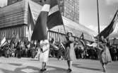 Första maj demonstration 2 maj 1967

Tre äldre kvinnor kommer gående i ett första-majtåg utanför varuhuset Krämaren i centrala Örebro. De tre kvinnorna bär en svensk flagga var i sina händer. De är klädda i ljusa kappor och mörka skor. Kvinnan i mitten har en ljus hatt på huvudet och kvinnan till höger har en sjalett på huvudet. I bakgrunden syns en folksamling. Fler fanor skymtar där som exempelvis amerikanska flaggan och israeliska flaggan.