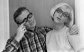 Glasögon 28 juli 1967

Närbild på en ljushårig kvinna med pagefrisyr som har ett par glasögon och en vit tröja på sig samt vita pärlörhängen som står tillsammans med en man klädd i rutig skjorta. Han håller sin vänstra arm om kvinnan. Med den högra handens pekfingar pekar han mot sina glasögon.