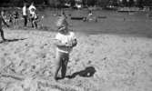 Grybe golf, Gustavsvik 17 juni 1967

En liten pojke i ettårsåldern klädd i en liten vit tröja och med bar underkropp står i förgrunden på utomhusbadet i Gustavsvik. Han håller i ett litet fat som han har fyllt med sand.