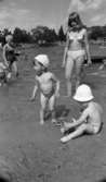 Grybe golf, Gustavsvik 17 juni 1967

I förgrunden syns två små pojkar i ettårsåldern som befinner sig i närheten av vattnet på utomhusbadet i Gustavsvik. Båda två har vita små mössor på sina huvuden och är i övrgt nakna. Pojken till vänster står upp. Det står en leksakshink fylld med sand och med en spade i vid hans fötter. En tonårsflicka står upp i bakgrunden i närheten av pojkarna och hon är klädd i en vit bikini. Bredvid henne på andra sidan sitter den andre lille pojken i sanden och håller en leksaksspade i sin högra hand. Framför honom står en leksakshink som han välter med vänstra handen. En kvinna i mörk baddräkt och en liten pojke i ljusa badbyxor syns i bakgrunden till vänster. Fler personer skymtar längre bort.