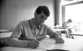Gymnasielgh, Fick körkort, Domuschefer, Socialhögskolan 9 juni 1967

En ung man klädd i grå tröja sitter vid ett skrivbord i ett klassrum och skriver på papper. I bakgrunden syns en kvinna som sitter och skriver vid ett annat skrivbord i närheten av ett fönster.