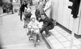 Gymnasielgh, Fick körkort, Domuschefer, Socialhögskolan 9 juni 1967

En liten flicka i sexårsåldern klädd i vit kappa med luva, svarta mönstrade strumpbyxor och mörka skor med vit rand och snöre framtill sitter på en trehjuling. På marken löper ett vitt streck. Hon är placerad vid detta. Ett litet trafikljus syns längre fram framför flickan. En man med mörk kavaj, ljusa byxor, ljusa strumpor och skor sitter på huk bredvid flickan. Två andra flickor sitter på rad på andra små fordon bakom den förstnämnda flickan. Längst bak i raden står en polisman. Andra barn och vuxna syns i bakgrunden.
