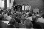 Göran Kummelstedt (Ej skannat), I3, Ekeskolan, Blombukett 13 juni 1967

En grupp ungdomar står på aulans scen på Ekeskolan i Örebro. Flickorna är klädda i klänningar och pojkarna är klädda i kavajer och kostymer. Nedanför scenen sitter publik i form av andra ungdomar och betraktar gruppen på scenen.
