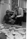 Göran Kummelstedt (Ej skannat), I3, Ekeskolan, Blombukett 13 juni 1967

En kvinna i en långärmad klänning sitter på golvet i ett rum och håller en bunt brev i vänsterhanden. Golvet framför henne är fyllt med brev. Bredvid henne sitter en man på huk stödd på sitt högra knä klädd i vit skjorta, svart slips, mörka byxor, mörka strumpor och mörka skor. Han tar ett brev med högerhanden ur kvinnans brevbunt.