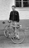 Palle Munther 18 maj 1967

En ung man klädd i svart sportjacka med ljusa kanter, rutig skjorta, grå byxor, svarta strumpor och vita sandaler står och håller i en sportcykel.