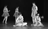 Göthman, Handikappfest,18 maj 1967

Fem kvinnor befinner sig på scenen under en revyföreställning på teatern. Alla är klädda i bikiniöverdelar, bastkjolar och har blomsterkransar runt halsarna. Två av dem sitter i förgrunden och spelar på trianglar. De tre andra står upp och dansar omkring och en av dessa kvinnor spelar också triangel.