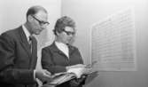 Länsläkaren, Trafiken 24 december 1966

En kvinna och en man står framför ett schema som hänger på en vägg i ett rum. Kvinnan är klädd i en ljus blus med kavaj över samt bär glasögon. Mannen bär också glasögon och är klädd i en grå kostym, vit skjorta och randig slips. De håller gemensamt i en uppslagen pärm. Båda har pennor i händerna och kvinnan pekar på schemat med sin penna.