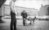 Med pappa på turné 14 april 1967

Den kände skådespelaren Helge Skoog spelar boule tillsammans med en annan man. Helges ettårsgamla barn sitter i en vit barnvagn med dubbelsits i närheten. De befinner sig på parkeringen bakom Norlings bryggeri. Helge är klädd i vit kavaj och svarta byxor. Hans barn är klädda i svarta dressar med svarta hattar på huvudena. Den andre mannen är klädd i svart jacka, randig skjorta, svarta byxor och svarta skor.