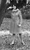 Modenummer 21 oktober 1966

Närbild på en ung kvinna som står och poserar invid ett träd. Hon är fotomodell. Hon är klädd i en ljus kappa med pälskrage och svarta, lågklackade skor.