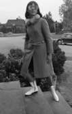Modenummer 21 oktober 1966

Närbild på en ung kvinna som går uppför en trappa och poserar samtidigt.Hon är fotomodell och är klädd i en ljus kappa med pälskrage och vita lågskaftade stövlar. Hon håller ett par ljusa handskar i sin högra hand. Hon har ljusa pappglasögon på sig.