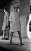Modevisning, Görtz motor (Rep av oil) 6 april 1967

Under en modevisning går en fotomodell över scenen. Hon är klädd i en vit kappa, vit hatt och har silverskor på fötterna. En skylt med delvis dold text hänger på väggen bakom henne. Till vänster i bakgrunden står en manlig fotomodell. Han är klädd i beige, rutig rock, beige byxor och mörka skor. Han har en mörk hatt på huvudet.