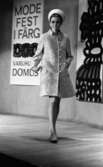 Modevisning, Görtz motor (Rep av oil) 6 april 1967

En fotomodell går på en scen under en modevisning. Hon är klädd i en kort, mönstrad ljus kappa, vita skor, vit hatt och har vita örhängen i öronen. Bakom henne syns en skylt på väggen där det står: 