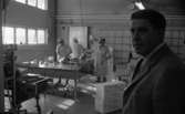 Lockhyttans herrgård, Kornbrödsbagaren, Varbergaskolan trivselskolan 17 februari 1968

Inne i fabriken Kornbrödsbagaren. I förgrunden står en man klädd i grå kostym, vit skjorta och mörkrandig slips. I bakgrunden syns kvinnor i vita arbetskläder och skyddsmössor som arbetar med bröd på ett bord.