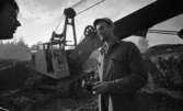 Vivalla Special Byggnummer 11 november 1967

Närbild på en arbetare i arbetsklädsel som håller i ett par hörselskydd. Han samtalar med en annan man. En grävmaskin syns i bakgrunden.