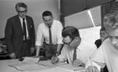 Mekanförbundet, 31 maj 1968

Två män sitter vid ett bord förgrunden och skriver på Mekanförbundet. De sitter med ritningar och skriver på papper och gör olika matematiska beräkningar. De går en mekanikerutbildning. De är klädda i vita skjortor och ljusa slipsar. Intill dem står en lärare i mekanik klädd i ljus skjorta ljus slips och ljusa byxor tillsammans med en annan herre som är klädd i mörk kostym, vit skjorta och mörk slips.