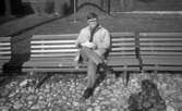 Wadköping (...?), 30 augusti 1968

En man sitter på en parkbänk i Wadköping. Han är med och ordnar eller repeterar en teaterföreställning. Han är klädd i beige rock, ljus skjorta, mörk slips, ljusa byxor, mörka strumpor och mörka skor. Han bär även glasögon samt håller några pappersark i sina händer. Två andra parkbänkar står på ömse sidor om honom. Byggnader syns i bakgrunden.