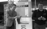 Korvätandet 30 augusti 1968

En man står och håller i en grillkorv som han precis har köpt i en automat. Han är klädd i ljus kavaj, ljus skjorta, mönstrad slips och mörka byxor. Bredvid honom står en man i mörk tröja och ljusa byxor. Han håller något i sina händer.