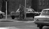 Vänstersväng med cykel 8 september 1967

En man klädd i beige rock och mörka byxor sitter på en cykel och har ena foten i gatan då han har stannat vid ett trafikljus på Rudbecksgatan i Örebro. Bilar passerar förbi och byggnader syns i bakgrunden.